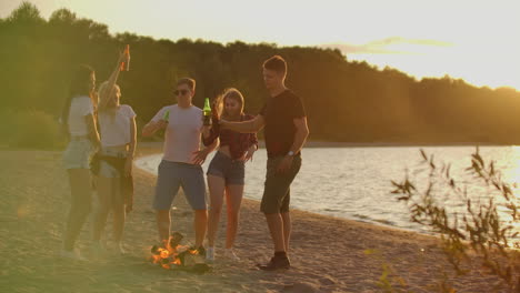 Eine-Gruppe-Von-Jungen-Und-Mädchen-In-Shorts-Und-T-Shirts-Tanzt-Mit-Bier-Um-Das-Lagerfeuer-Am-Sandstrand.-Sie-Genießen-Den-Sommerabend-Am-Flussufer-Bei-Einer-Open-Air-Party.